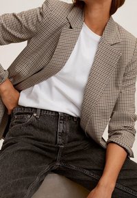 Personne portant un blazer à carreaux beige sur un t-shirt blanc et un jean noir délavé taille haute, la main posée sur la hanche.