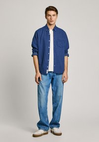 Blue patterned button-up shirt over a white tee, paired with light blue denim jeans and white sneakers with brown accents.