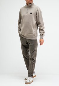 Grauer Halbzip-Pullover mit kleinem Logo, kombiniert mit olivgrünen Hosen und weißen Sneakers mit grauen Akzenten. Glattes Material und entspannte Passform.