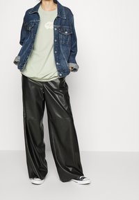 Giacca di jeans blu scuro, indossata sopra una maglietta verde chiaro con logo, abbinata a pantaloni di pelle nera larghi e sneakers nere.