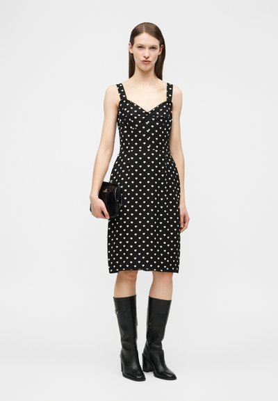 Γυναίκα που φοράει μαύρο φόρεμα μέχρι το γόνατο με λευκές καρδιές, κρατώντας μαύρο φάκελο και φοράει μαύρες ψηλοτάκουνες μπότες μέχρι το γόνατο, στέκεται μπροστά σε λευκό φόντο.