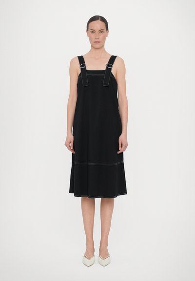 Schwarzes Kleid mit breiten Trägern, silbernen Beschlägen und weißen Nähten. A-Linie mit einem ausgestellten Saum. Getragen mit weißen High Heels.