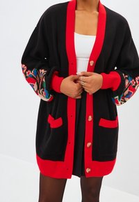 Black cardigan with red collar and pockets, featuring colorful floral embroidery on sleeves. Five buttons down the front. Soft texture.