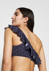 Marineblå bikini topp med prikkete mønster og en skulderdesign, med volangdetalj og glatt tekstur. Bakvisning som viser minimal dekning.