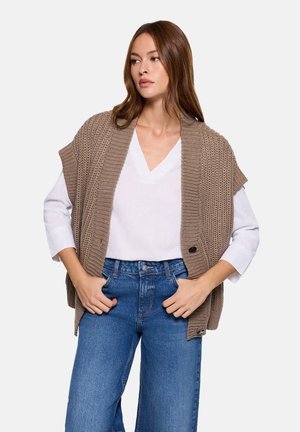 Chaleco beige de punto con cuello ancho tipo chal, patrón texturizado y botones, llevado sobre una camiseta blanca de escote en V y jeans azules de talle alto.