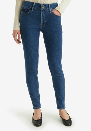 Vrouw draagt blauwe skinny jeans, een crèmekleurige geribde trui en zwarte ballerina’s, met één hand in de zak en de andere ontspannen.