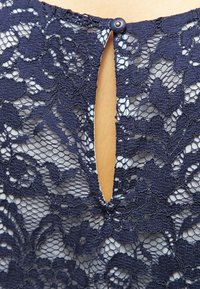 Haut en dentelle marine avec un motif floral, encolure dégagée avec un détail en forme de trou de serrure, et fermeture par bouton dans le dos. Texture en dentelle évidente.