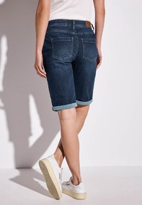 Jeansshorts i mörkblått med uppvikt fåll, som har två bakfickor och en brun läderpatch vid midjan. Vita sneakers fulländar looken.