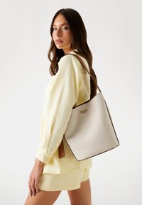Beige tote bag med struktureret stof, brune læderdetaljer og logo. Sammen med en lys gul linned bluse og shorts.