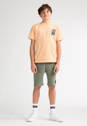 T-shirt en coton pêche clair à manches courtes avec un motif noir sur la poitrine, associé à un short vert olive, des baskets blanches et des chaussettes à rayures noires.