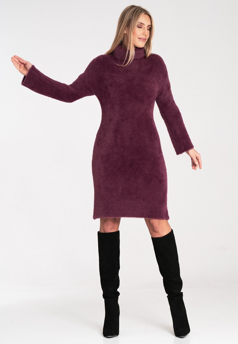 Pulloverkleid mit langen Ärmeln in tiefem Lila, strukturiertem Stoff, figurbetontem Schnitt, endet oberhalb des Knies, kombiniert mit schwarzen knielangen Stiefeln.