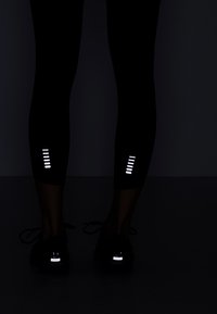 Černé legíny s reflexními akcenty na spodní části nohavic. Texturovaná tkanina, úzký střih, spárované s černými sportovními botami s reflexními prvky.