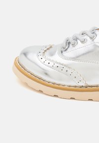 Scarpa in pelle argentata con dettagli brogue, punta arrotondata e suola in gomma beige. Presenta perforazioni decorative e lacci tonali.