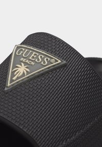 Κοντινό πλάνο του ιμάντα ενός μαύρου σαγιονάρου με ανάγλυφη υφή, με το μπεζ τρίγωνο λογότυπο Guess Beach και το έμβλημα με φοίνικες.