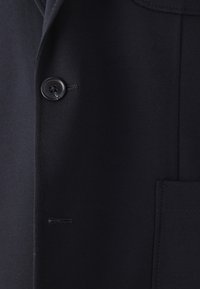 Nærbillede af en marineblå blazer, der viser en sort knap med "Marc O'Polo" indgravering, en knaphul og en syet lomme på højre side.