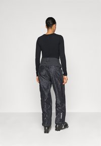 Top preto de manga longa com decote redondo, combinado com calças cargo de padrão escuro e botas de ski pretas, visto por trás.