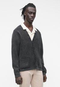 Anglies spalvos rūbų audinio megztinis su bangelėmis, turintis V formos iškirptę, du priekinės dalies kišenes ir sagų užsegimą, dėvimas ant dryžuotos marškinių.