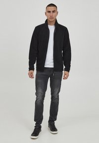 Chaqueta de forro polar negra con un cuello alto, cierre de cremallera, llevada sobre una camiseta blanca. Combinada con jeans gris oscuro y zapatillas negras.
