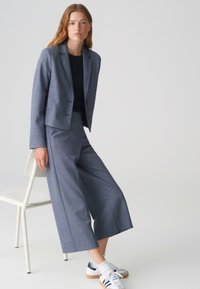 Blazer sur mesure gris avec un pantalon large assorti, associé à des baskets blanches. La tenue présente une texture douce et un design minimaliste.