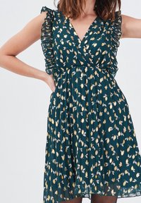 Robe florale verte avec un décolleté en V profond et des manches volantées. Caractérisée par un motif de feuilles dorées, une jupe plissée et un tissu léger et fluide.