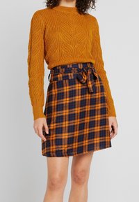 Pull en tricot orange avec un motif texturé, assorti à une jupe à carreaux dans des nuances de bleu marine et orange, avec une ceinture nouée à la taille.