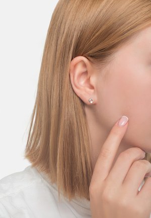 Silberne Ohrstecker mit kreisförmigem Design, ausgestattet mit einer glatten Metalloberfläche. Das Model zeigt einen Ohrstecker und berührt dabei ihre Wange.