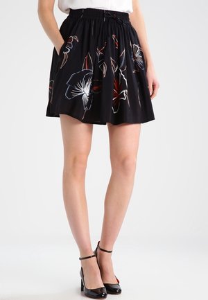 Frau, die einen schwarzen Blumenrock mit elastischem Bund und schwarzen Riemchensandalen trägt, steht mit den Händen in den Rocktaschen.