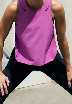 Persoon met een mouwloos roze sporttop en zwarte leggings, voorovergebogen met handen op de knieën, buiten in fel daglicht.