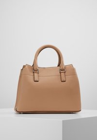 Tan pebblat läderhandväska med dubbla rundade handtag, strukturerad form och silverfärgad hårdvara, uppställd på en vit pedestal mot en ljusgrå bakgrund.