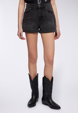 Shorts di jeans neri a vita alta con risvolti, dotati di passanti per cintura e tasche frontali. Abbinati a stivali cowboy neri.