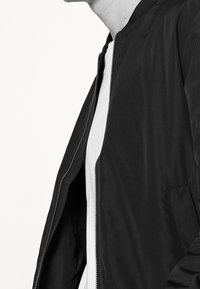 Černá bomber bunda se zipovým zapínáním, žebrovaným límcem, hladkou texturou a bočními kapsami, nošená přes bílou košili.