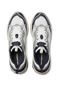 Balta un tumši zilā sporta apavi ar teksturētu virsmu, pelēkiem akcentiem, noapaļotu formu un plakaniem šņorējumiem. Atslēgas elements ir cilpiņa uz papēža.