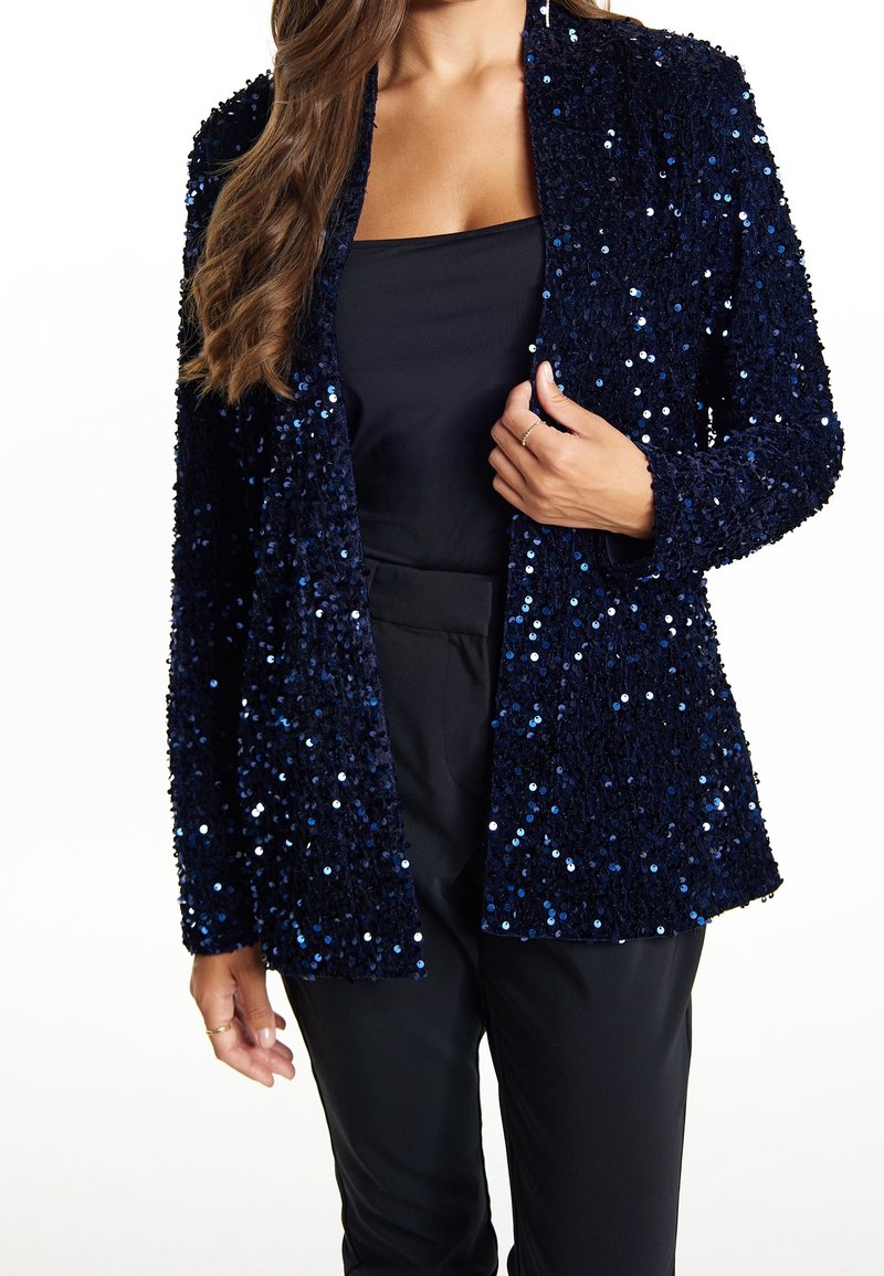 Blazer de lentejuelas en tono marino con una superficie texturizada, abertura frontal y mangas largas. Combinado con una blusa negra y pantalones negros.