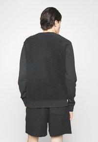 Man med mörkt hår som bär en svart långärmad sweatshirt och svarta shorts, stående vänd bort från kameran mot en vit bakgrund.