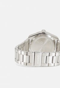 Reloj de acero inoxidable plateado con una pulsera brillante y enlazada y una caja redonda, con un dial minimalista y un logo grabado en la parte posterior.