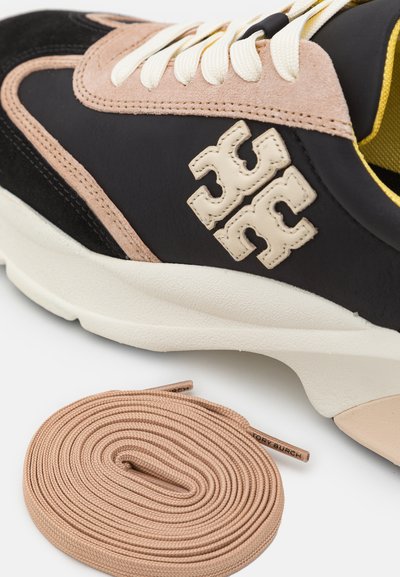 Svart och beige sneaker med mocka och lädermaterial, utrustad med tjock vit sula, kontrasterande gul kant och beiga snören.