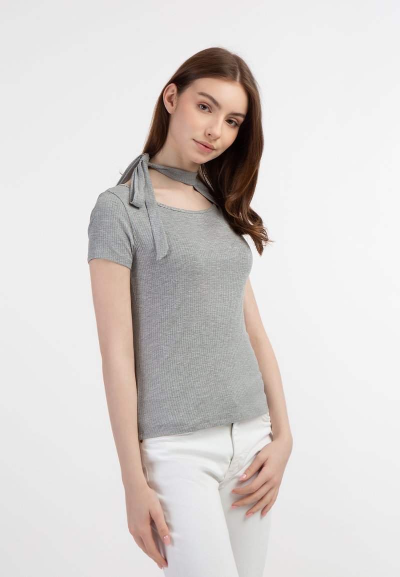 Lys grå ribbede t-shirt med korte ærmer, der har et choker-lignende halsbånd og en tætsiddende pasform. Sammen med hvide bukser.