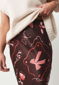 Jupe marron présentant un motif de nœuds roses, de cocktails et de motifs festifs. Tissu lisse avec une finition brillante. Taille élastique visible.