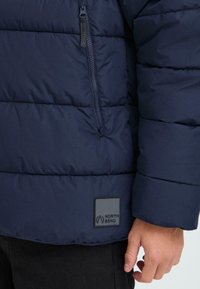 Granatowa kurtka puchowa z zamek błyskawicznym, z kieszenią zapinaną na zamek, z kwadratową naszywką z napisem "NORTH BEND" z boku. Miękka, pikowana faktura.