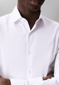 Hvid skjorte med knapper og klassisk krave, med en glat tekstur og et diskret logo på brystet, båret med krydsede arme.