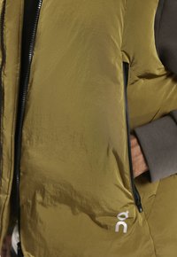 Person in senfgelber gepolsterter Weste und grauem Ärmelkleidungsstück, Hand ruht in der seitlichen Reißverschlusstasche der Weste.