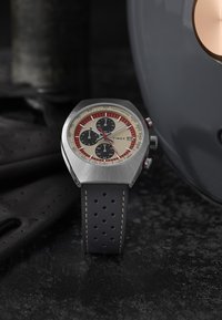 Montre Timex avec un boîtier en acier inoxydable, un cadran beige, des sous-cadrans noirs, une lunette tachymétrique rouge et un bracelet en cuir perforé noir.
