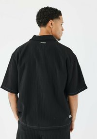 Zwarte short-sleeve overhemd met een gestructureerd oppervlak, open kraag en witte stikdetails. Beschikt over een logo op de bovenrug en heeft een losse pasvorm.