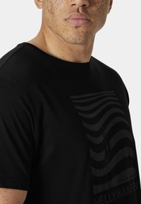 Camiseta negra de algodón con cuello redondo, que presenta un patrón de olas en tono y el logo de la marca en la parte delantera. Textura suave y diseño casual.
