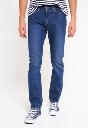 Mężczyzna ubrany w niebieskie dżinsy, granatowe trampki z białymi sznurowadłami i pasiastą koszulę, stojący na białym tle.