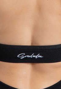 Zwarte geribbelde sport beha met een zachte elastische band, voorzien van een witte cursieve logo op de achterkant. Fluweelachtige textuur, minimalistisch ontwerp.