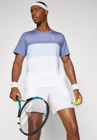 Lys blå og hvit tennisgenser med horisontale striper, sammen med hvite shorts. Har en logo på brystet og en tennisracket i hånden.
