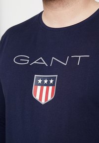 Navyblaues Baumwoll-T-Shirt mit einem großen "GANT"-Logo in Grau und einer Schildgrafik mit drei Sternen und roten Streifen. Rundhalsausschnitt.