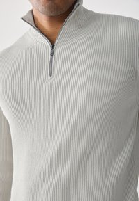 Suéter de canalé gris claro con cuello de cremallera de un cuarto. Tela suave y patrón texturizado, con una cremallera cerca del collar para un ajuste ajustable.