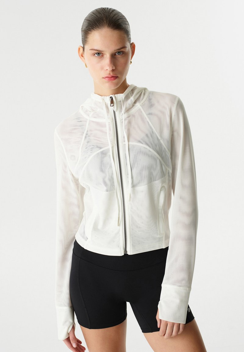 Veste zippée blanche avec une capuche, fabriquée en matériau transparent, dotée de manches longues et de bords côtelés, associée à un short noir.